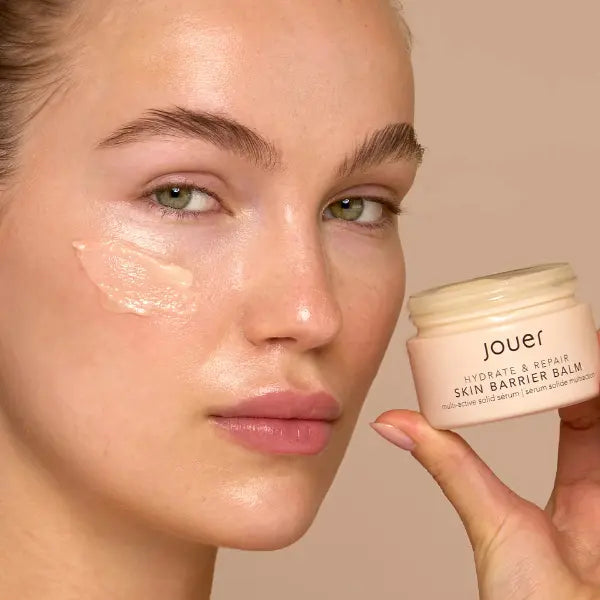 Antioxidant skincare product applied to model's cheek with Jouer Skin Barrier Balm jar held beside her face.
