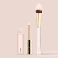 Liquid concealer and brush set in Snow, a very light neutral shade with pink undertones, featuring a doe-foot applicator and tapered blending brush on a beige background.