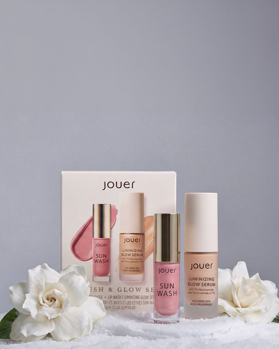 Jouer Blush & Glow Set featuring Sun Wash Lip + Cheek and Luminizing Glow Serum, displayed with white gardenias on a snowy holiday backdrop.