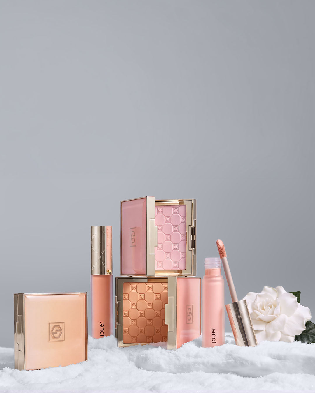 Jouer makeup collection featuring blush, bronzer, face powders, and lip products in pink and gold packaging, surrounded by snow and white flowers.
