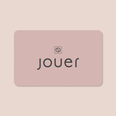 The Gift Card from Jouer Cosmetics features a rectangular design in a soft, light pink with rounded corners. "jouer" is displayed in lowercase black letters centered on the card, and above it sits a small black icon resembling a stylized, symmetrical letter "J" enclosed within a square. The background of the card is a gentle light beige.