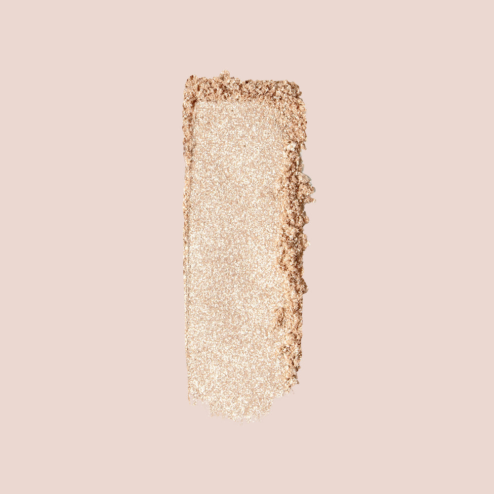 A smear of Jouer Cosmetics' Powder Highlighter in a shimmering, light golden beige hue graces a neutral background, showcasing its textured and powdery finish with a hint of sparkle. This cruelty-free highlighter is both vegan and paraben-free.