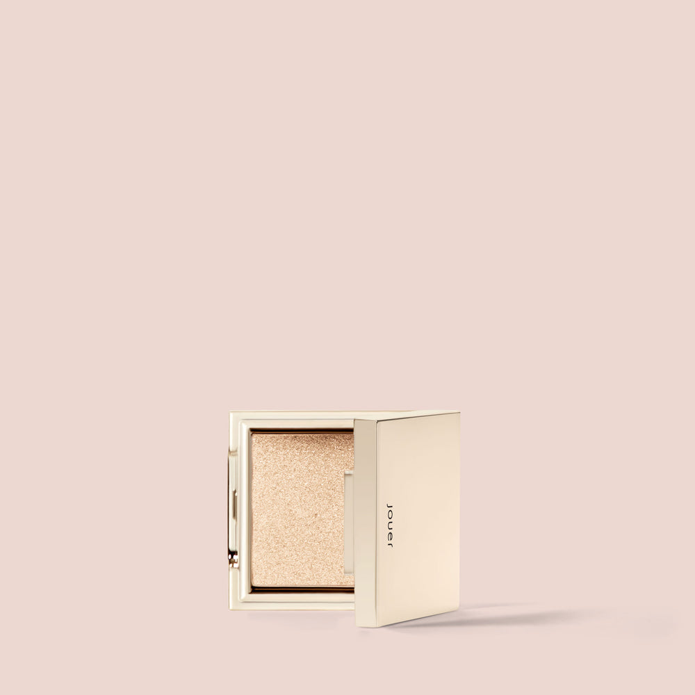 A sleek, square compact of Jouer Cosmetics' Powder Highlighter in a shimmering champagne hue is partially open against a plain beige background. This cruelty-free and vegan compact epitomizes minimalist elegance with its smooth edges and subtle shine.