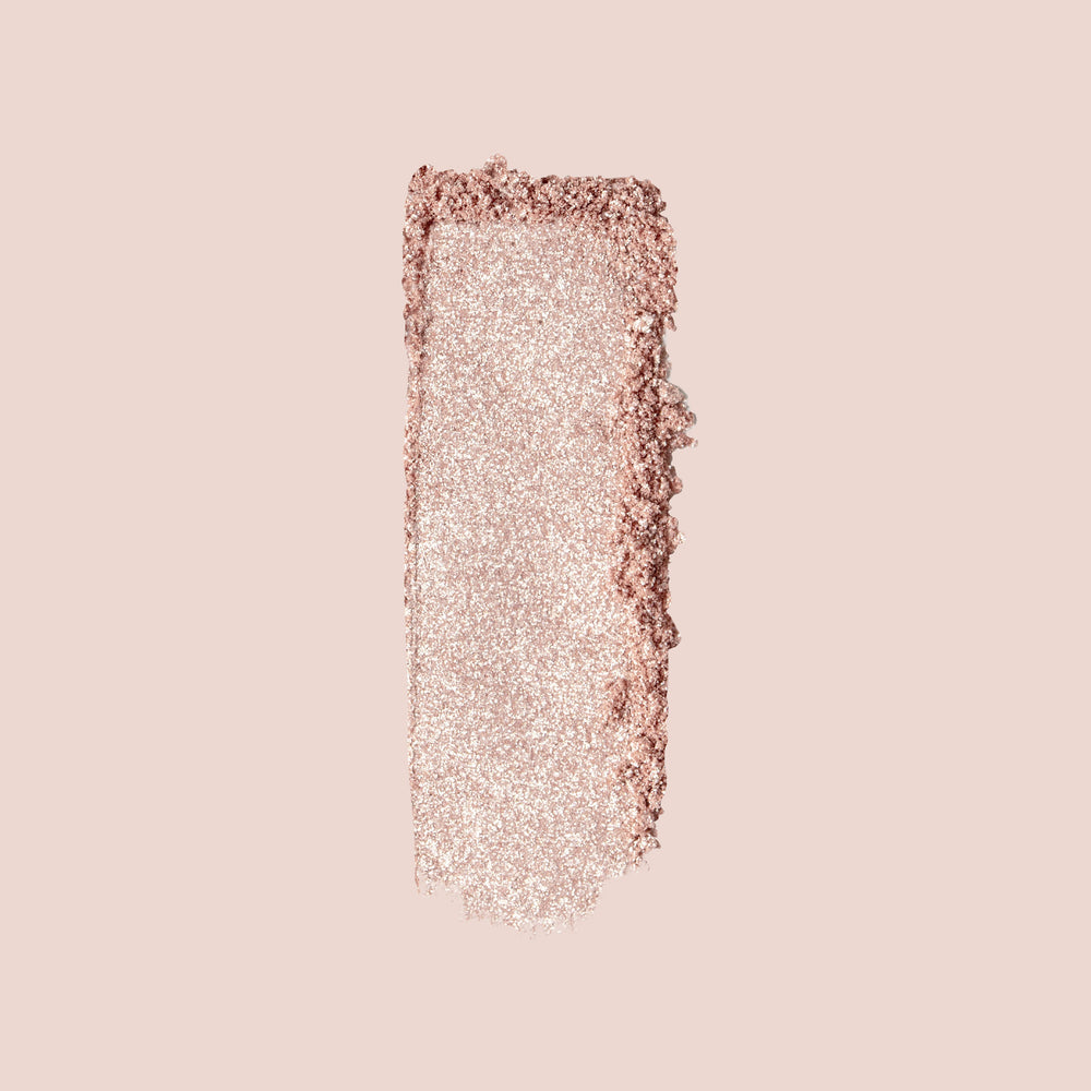 A dense, rose-gold rectangular swatch of Jouer Cosmetics' Powder Highlighter is shown on a light beige background, highlighting its shimmering, vegan formula with sparkly texture and rich pigmentation.