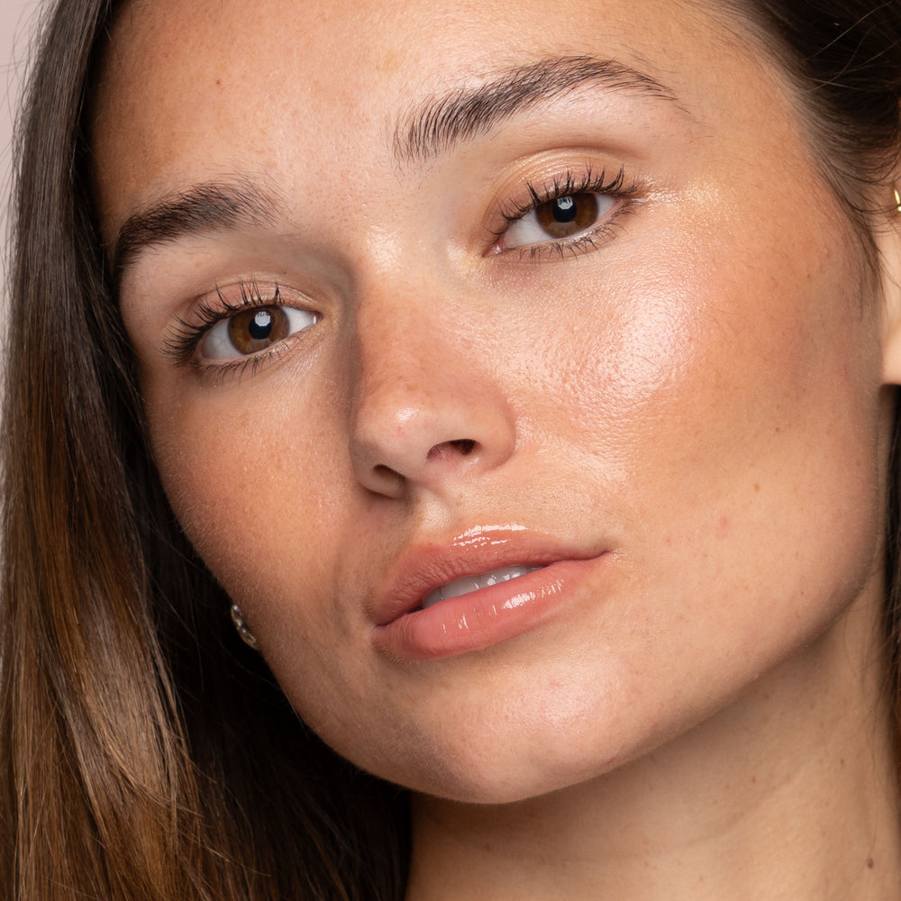 Close-up of a person with long brown hair and smooth, glowing skin. They have brown eyes, natural makeup, glossy lips, and are looking directly at the camera with a neutral expression. Their beauty routine features Jouer Cosmetics' Powder Highlighter to maintain such flawless, radiant skin using cruelty-free products.