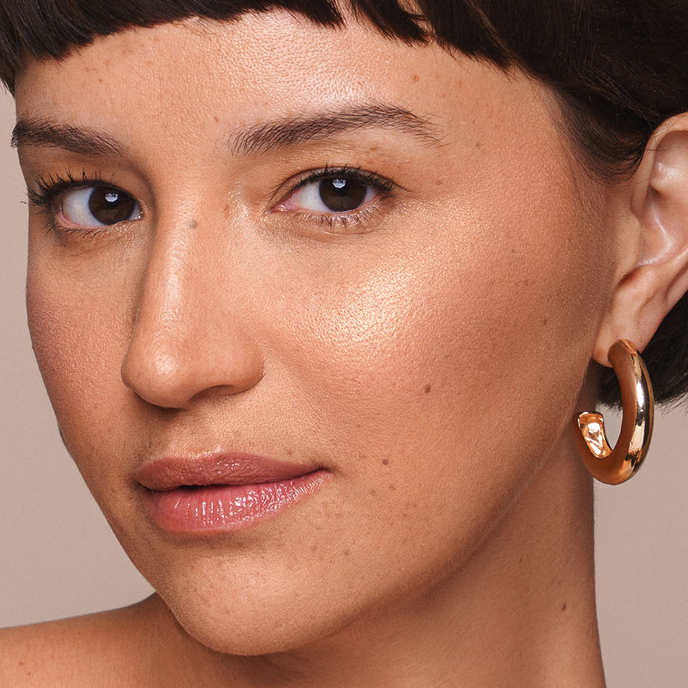 Close-up of a person with short dark hair, wearing large gold hoop earrings. Their skin has a natural glow, accentuated by the Powder Highlighter from Jouer Cosmetics. Their makeup includes subtle lip gloss and minimal eye makeup that is both vegan and cruelty-free. The background is plain and neutral, highlighting their facial features.