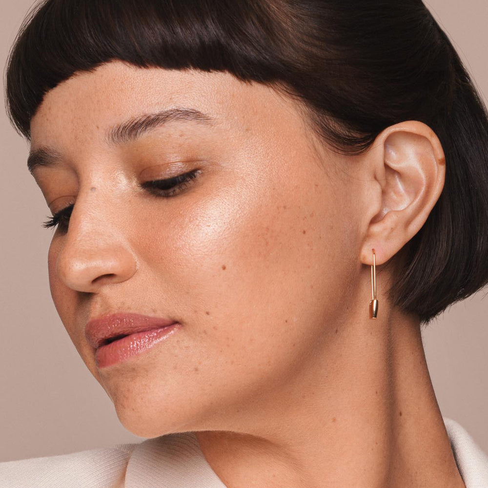 A person with short, dark hair and bangs showcases a minimalist gold earring. The close-up highlights dewy, glowing skin enhanced by Jouer Cosmetics' Powder Highlighter, a natural and vegan makeup product. The background is a neutral, light brown color, adding to the soft and elegant look of the image.