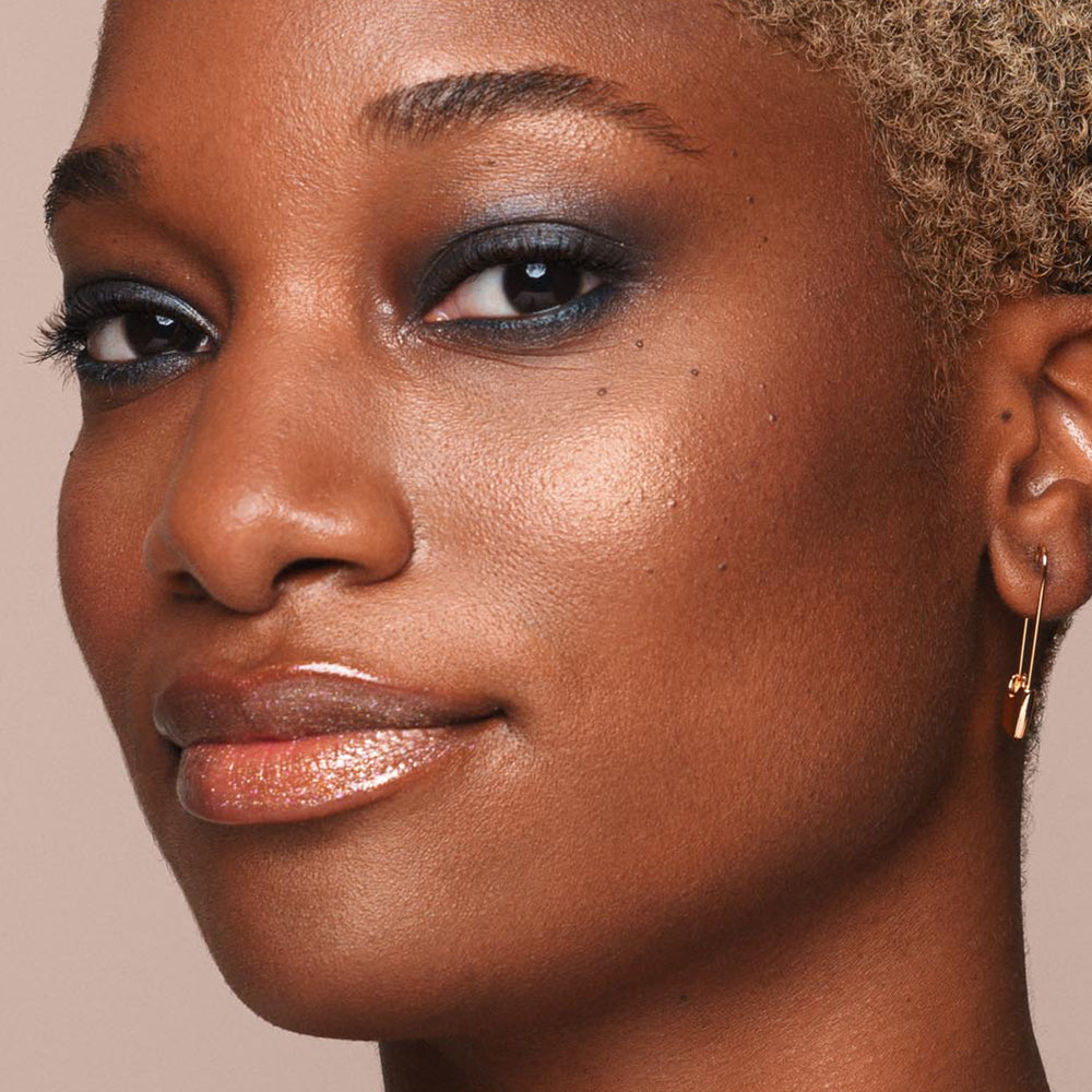 Close-up of a person with short, curly hair who has glowing skin, wearing shimmery eye shadow, glossy lip makeup, and small hoop earrings with a charm. The background is neutral, enhancing the focus on their radiant complexion accentuated by Jouer Cosmetics' Powder Highlighter, which is both vegan and cruelty-free.