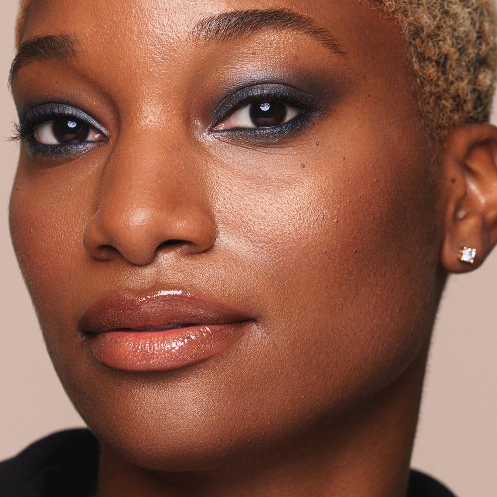 A close-up of a person with short blonde hair reveals their glistening skin, enhanced by Jouer Cosmetics' Powder Highlighter. Their glossy lips complement the smoky eye makeup in shades of black and gray. A small stud earring adorns one ear, while the neutral background accentuates their radiant face. All makeup used is cruelty-free and vegan from Jouer Cosmetics.