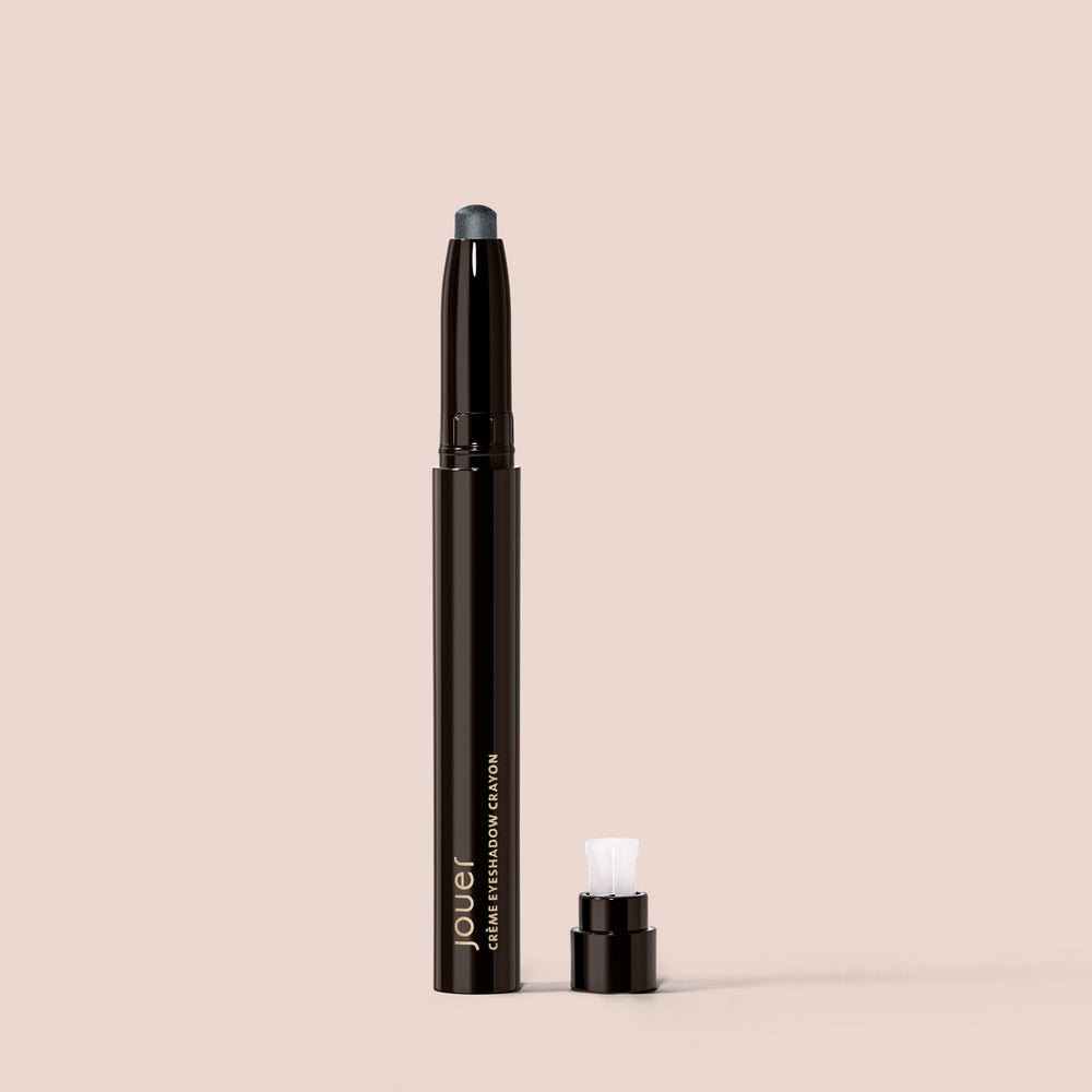 A Crème Eyeshadow pencil in Marble from Jouer Cosmetics, featuring a detachable cap, is set against a beige background. This cruelty-free pencil has a dark tip designed to define your eyelid, and it includes a white applicator positioned near the base next to the cap.