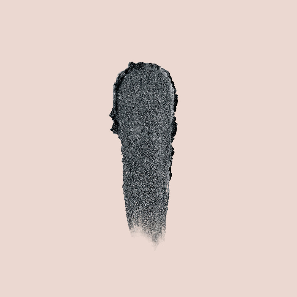 A vertical smear of Jouer Cosmetics' Crème Eyeshadow in a dark gray, textured finish on a beige background, resembling a stroke of cruelty-free paint or charcoal.