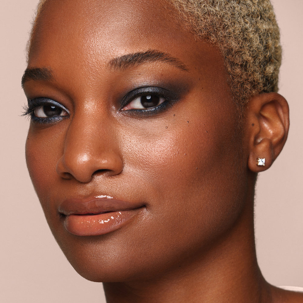 A person with short blonde hair, glossy lips, and dramatic dark eye makeup from Jouer Cosmetics' Crème Eyeshadow line looks directly at the camera. Flaunting a paraben-free radiant complexion and cruelty-free eyelid beauty, they wear a diamond stud earring against a neutral background.
