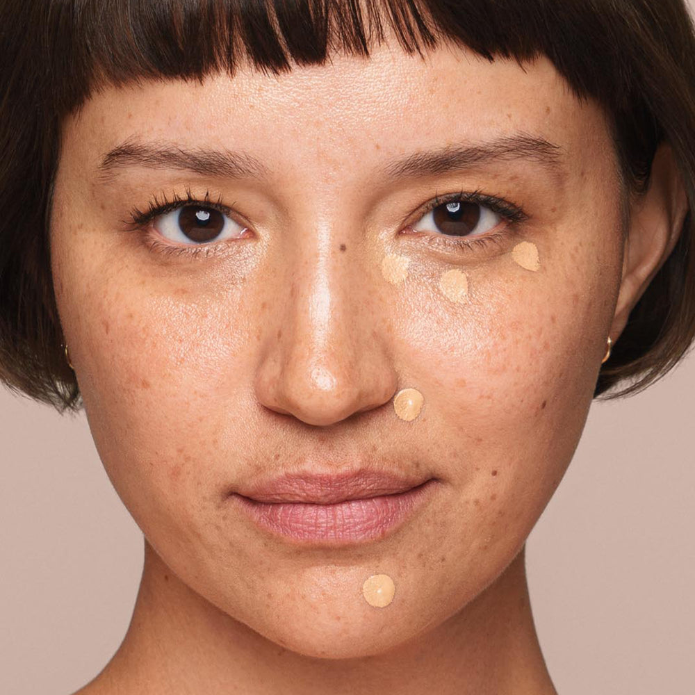 A person with short hair and freckles uses a concealer brush to apply dots of Jouer Cosmetics' Essential Liquid Concealer, which is paraben-free, under their eyes and on their chin. The background is plain and neutral-colored.
