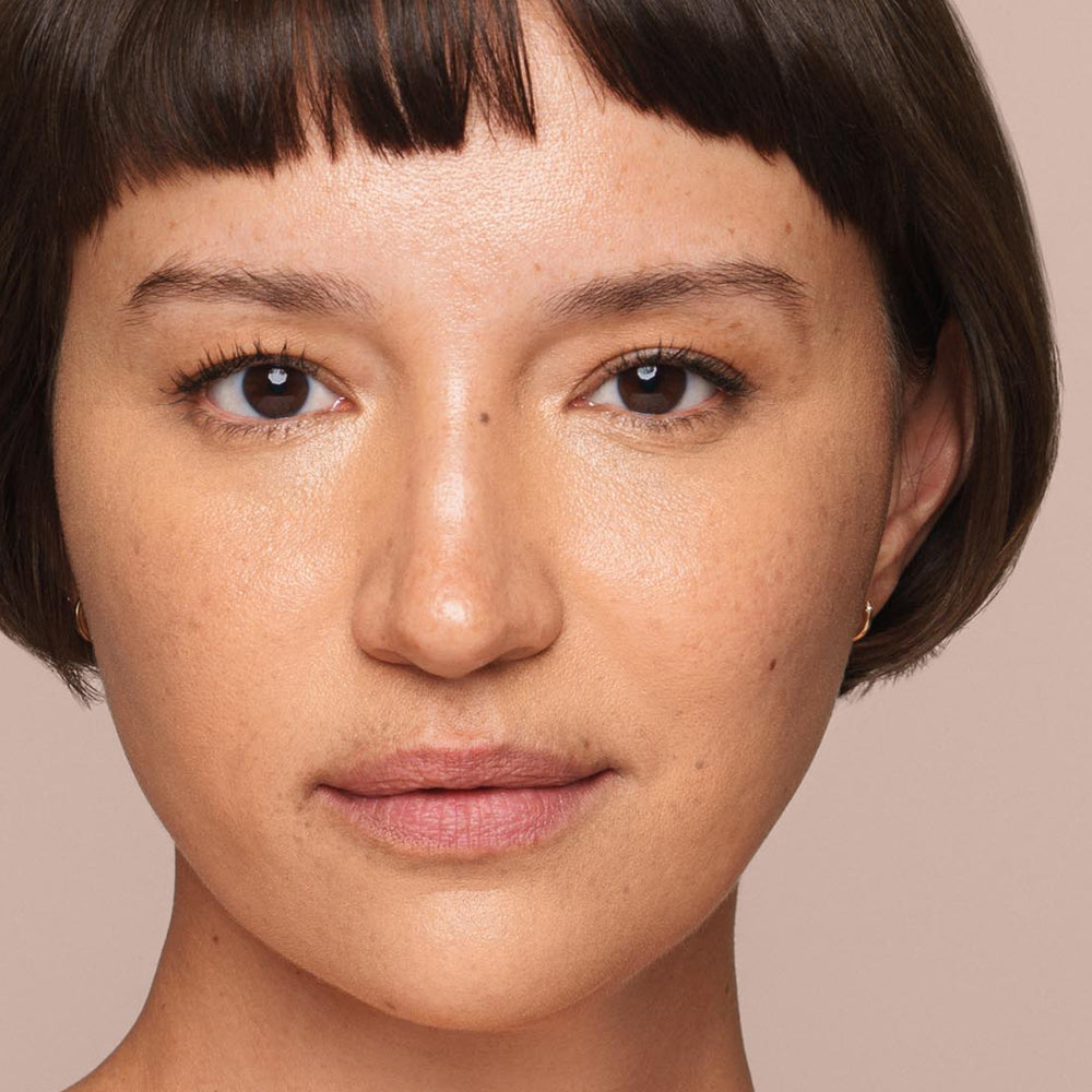 A close-up view of a person with short, dark hair, straight bangs, and brown eyes. The individual has a neutral expression, with minimal makeup applied using the Jouer Cosmetics Essential Liquid Concealer brush, highlighting natural skin texture and features. The background is a plain, light beige color.