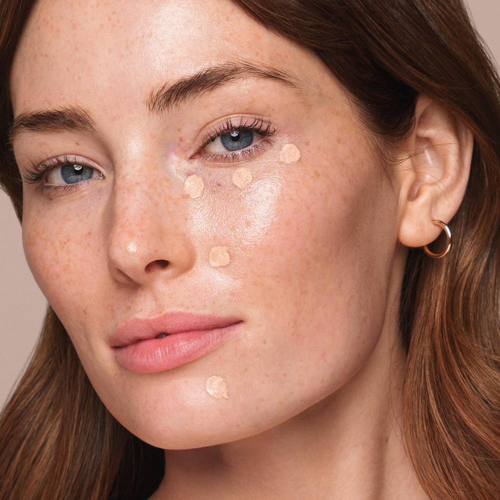 A close-up of a person with light skin, blue eyes, and freckles applying spots of Jouer Cosmetics' Essential Liquid Concealer under their left eye and on the left side of their face. They have natural-looking makeup, pink lips, and are wearing a small hoop earring.