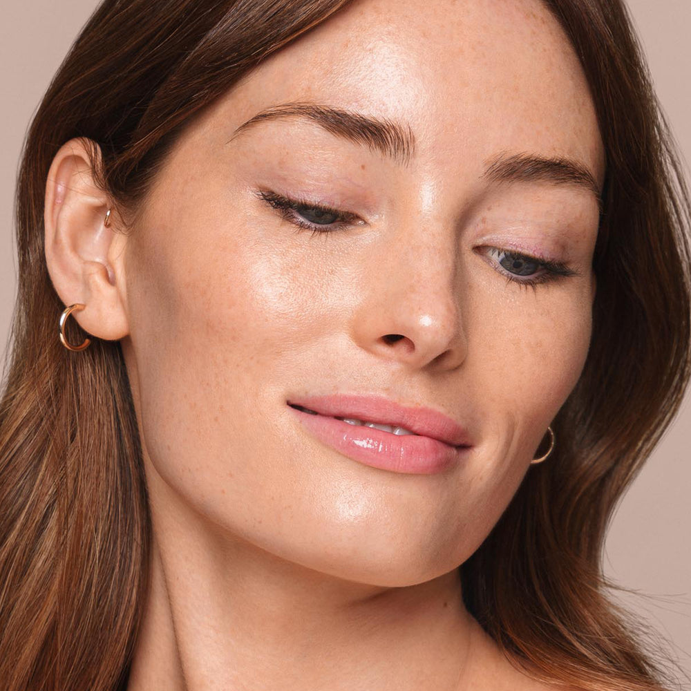 A woman with long brown hair and fair skin is looking down and slightly smiling. She is wearing small, gold hoop earrings and has minimal makeup, giving her a natural look that was likely achieved with Jouer Cosmetics' *Essential Liquid Concealer*. Her smooth skin has a slight glow against the beige background.
