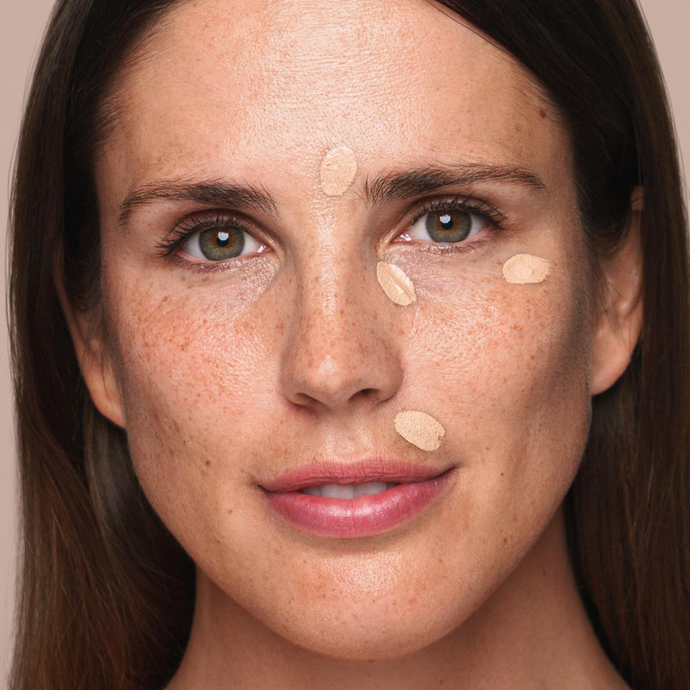 A close-up of a person with fair skin and brown hair, displaying a neutral expression. Small dabs of Jouer Cosmetics' *Essential Liquid Concealer are on their forehead, under their eyes, and on their cheeks, presumably in the process of being blended with a concealer brush. The background is plain cream.
