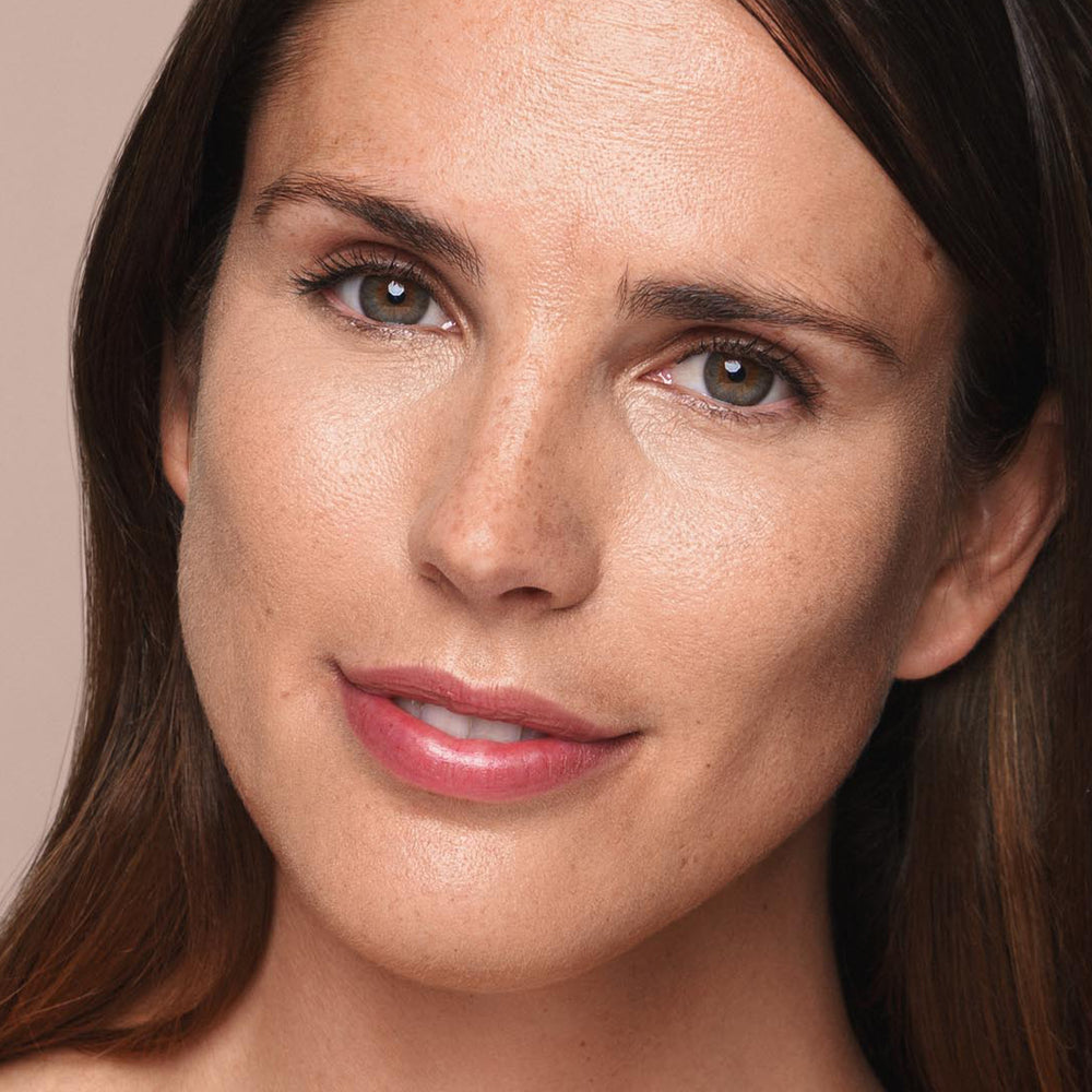 Close-up of a person with light skin, straight dark brown hair, and brown eyes. They have a neutral expression, slightly smiling, showing smooth skin with minimal makeup applied using Jouer Cosmetics' *Essential Liquid Concealer*. Their light pink lip color is complemented by vegan and paraben-free products against the plain beige background.