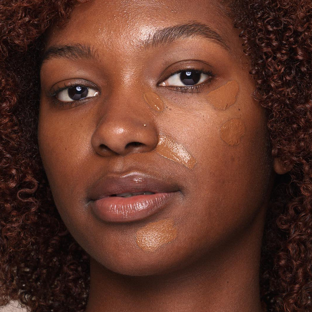 A person with deep brown skin and curly hair is shown up-close with natural makeup. Several swatches of Jouer Cosmetics' *Essential Liquid Concealer, which is paraben-free and slightly lighter than their skin tone, are applied to their cheek using a concealer brush. The person has a neutral expression.