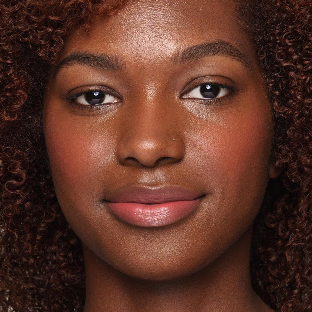 Close-up image of a person with a warm brown complexion, curly hair, and subtle makeup, using an *Essential Liquid Concealer* brush by Jouer Cosmetics to achieve natural eyebrows, light pink blush, and light pink lipstick. The individual has a small nose piercing on their left nostril and a serene, gentle expression.