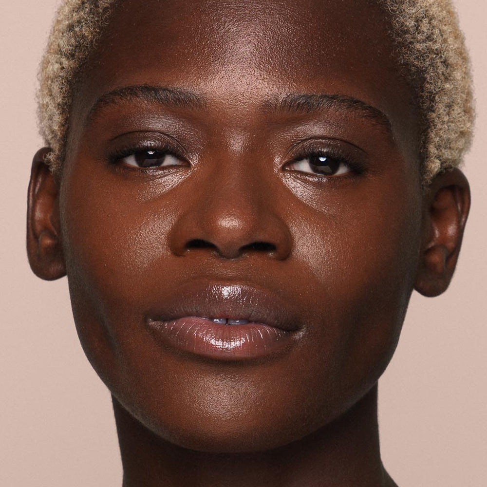 Close-up of a person with short, light-colored hair and a smooth complexion, looking directly at the camera. Their impeccably even skin speaks volumes about the effectiveness of Jouer Cosmetics' *Essential Liquid Concealer*, which boasts a paraben-free formula. The background is a plain, neutral beige color.