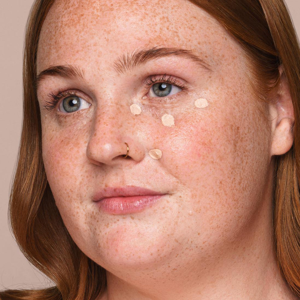 A person with light skin and freckles looks into the distance while applying several dots of Jouer Cosmetics' *Essential Liquid Concealer* under their eye on their left cheek. They have long, reddish-brown hair and a nose piercing, using a concealer brush in front of a neutral background.