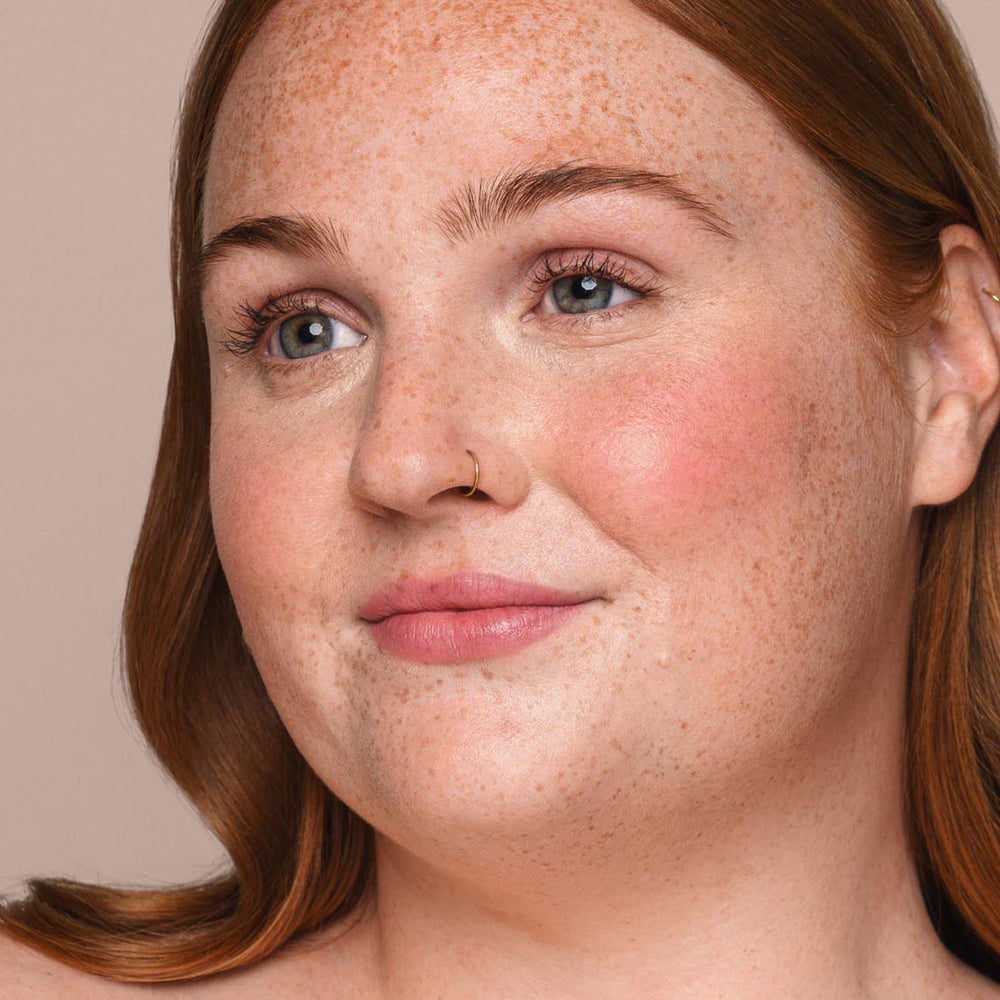 Close-up of a woman with light skin, blue eyes, and freckles, looking slightly to the side and smiling softly. She has long auburn hair, a nose ring, and is wearing light makeup featuring Jouer Cosmetics' Essential Liquid Concealer for a paraben-free natural look. The background is a neutral beige color.