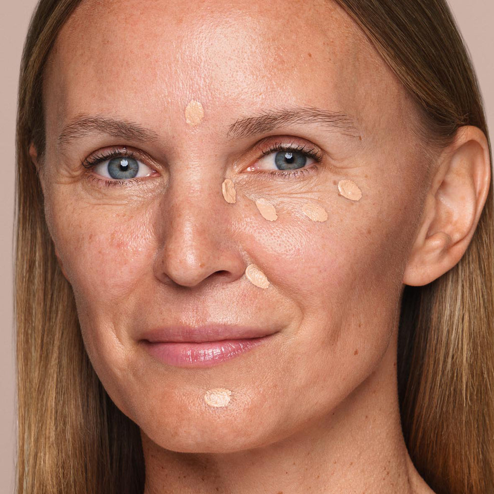 A woman with light skin and straight, light brown hair is shown with dots of Jouer Cosmetics' Essential Liquid Concealer on her forehead, under eyes, and on nose and cheeks, suggesting she is about to blend it with a concealer brush. She has a slight smile.