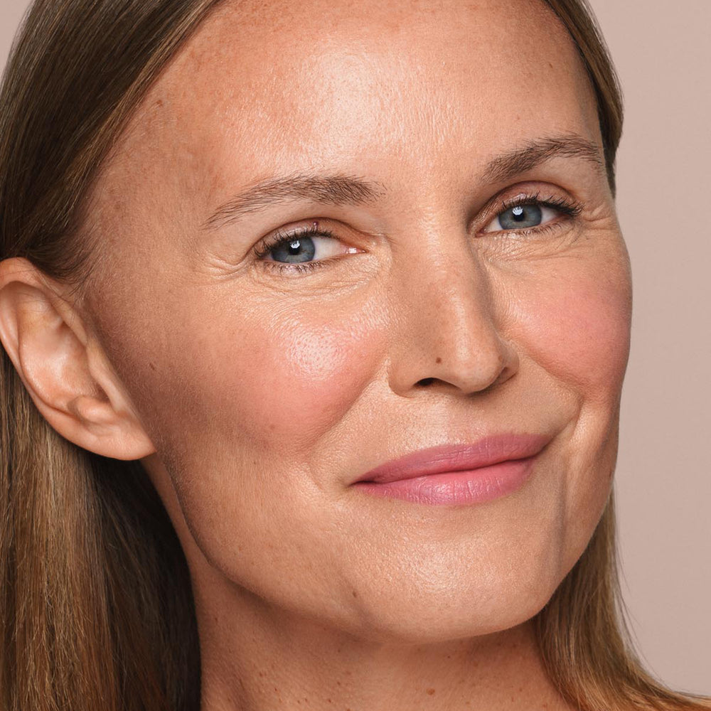 Close-up of a middle-aged woman with fair skin and blue eyes, smiling gently. She has straight, light brown hair worn down. The background is a plain, light beige color. The woman has a few fine lines and a natural look, achieved using vegan and paraben-free products with minimal makeup application using the *Essential Liquid Concealer* by Jouer Cosmetics with a concealer brush.