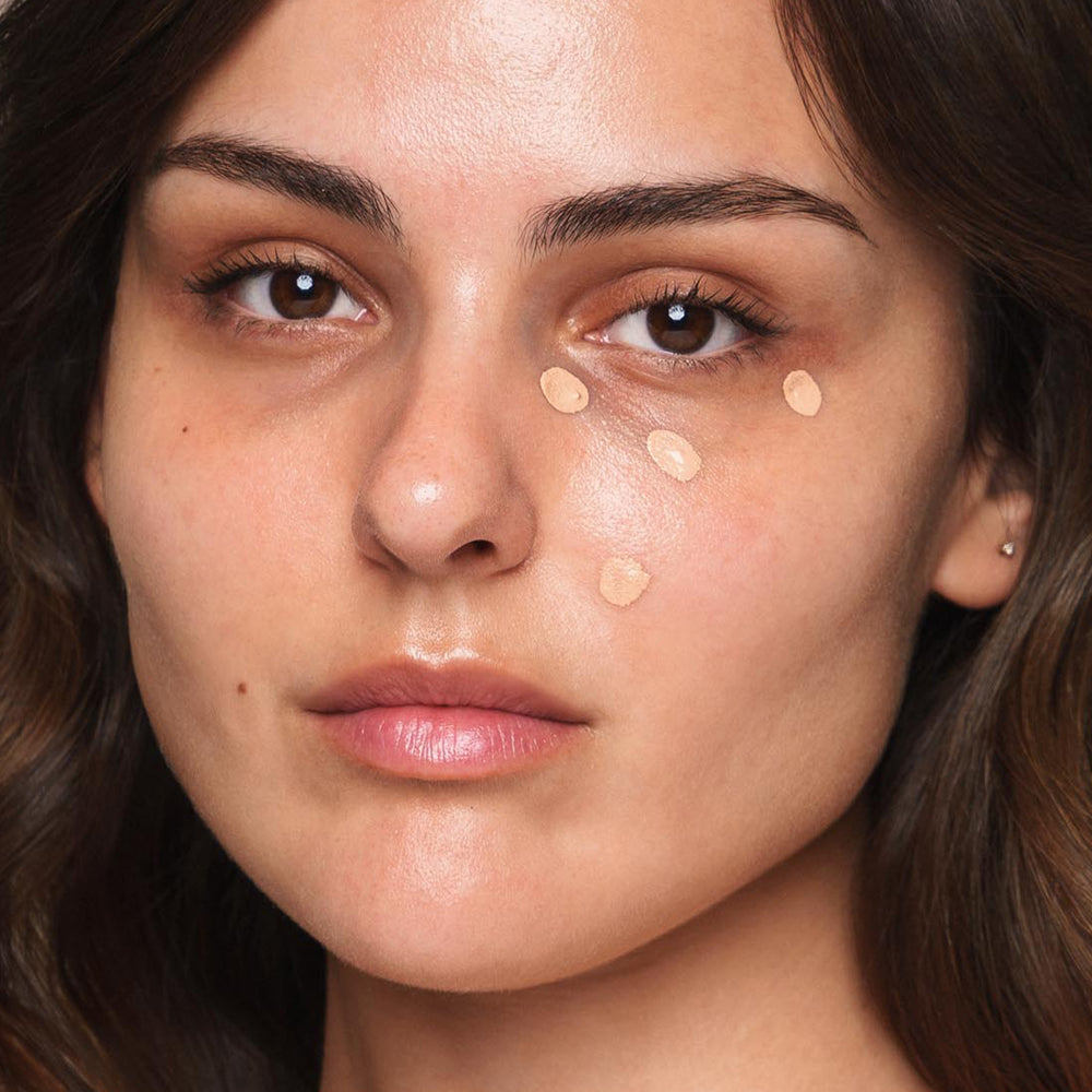 A close-up of a woman applying small dots of Jouer Cosmetics' Essential Liquid Concealer, which is vegan and paraben-free, under her eyes. She has brown hair and a neutral expression, with no makeup on the rest of her face.
