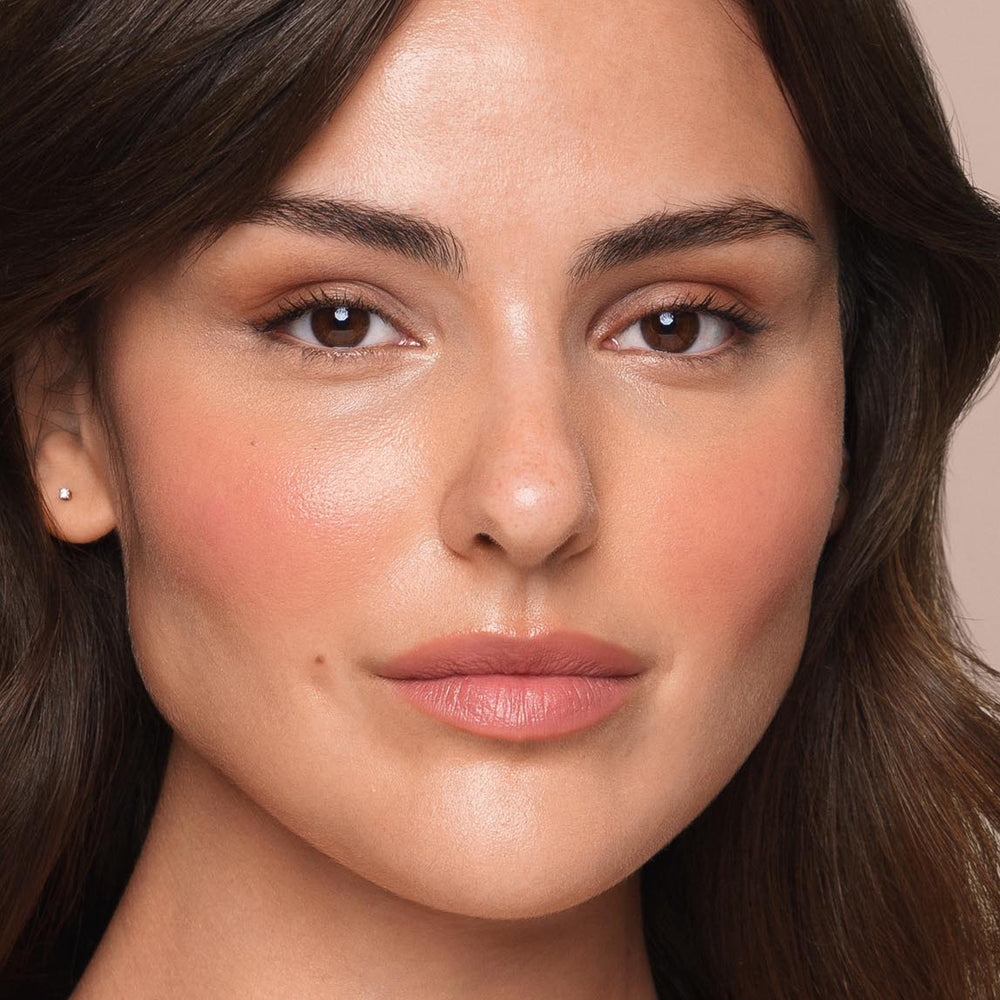 A close-up of a person with clear skin, minimal makeup, brown eyes, and brown hair. They are using the *Essential Liquid Concealer* by Jouer Cosmetics with a vegan concealer brush, looking directly at the camera with a neutral facial expression. Their cheeks have a subtle blush and they are wearing natural-colored lipstick.