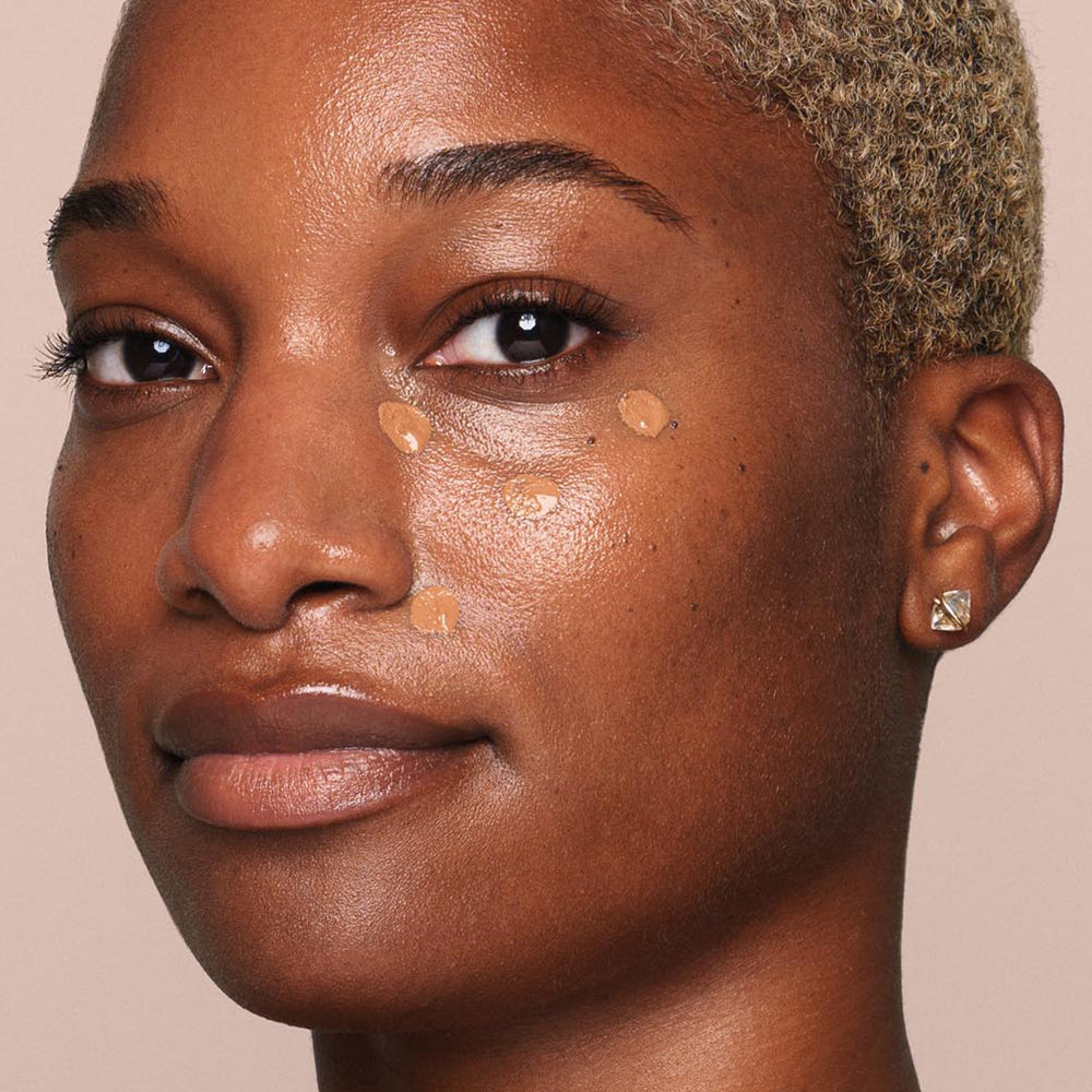 A person with short, blonde hair and a neutral expression looks at the camera with several small dots of Jouer Cosmetics' Essential Liquid Concealer applied under their right eye using a concealer brush. They wear a small, square-shaped earring in their right ear against a plain beige background.