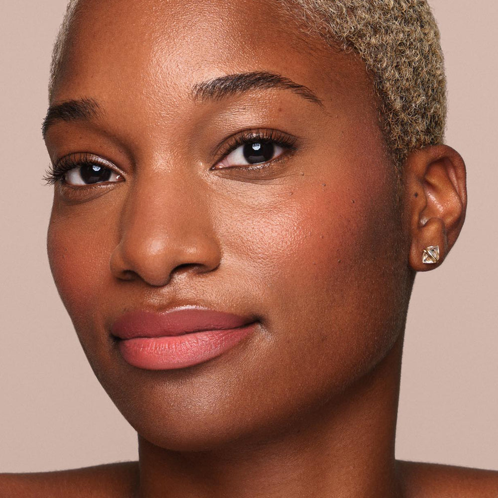 Close-up portrait of a person with short blonde hair and glowing skin, using paraben-free cosmetics from Jouer Cosmetics. They have subtle makeup, including pink lipstick and blush applied with the Essential Liquid Concealer brush, and are wearing small stud earrings. They are looking directly at the camera with a slight smile.