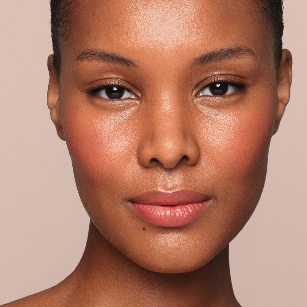 A close-up portrait of a person with smooth, glowing brown skin, wearing natural, vegan makeup. The person has slightly rosy cheeks, full lips with a hint of gloss, and minimal eye makeup that enhances their eyes. A meticulously applied Jouer Cosmetics Essential Liquid Concealer perfects the look against a neutral beige background.