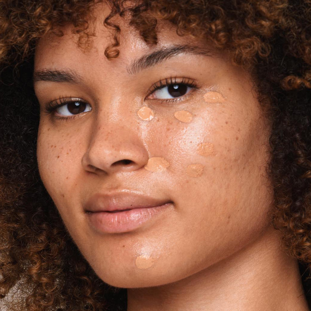 A close-up shows a person with medium skin tone, curly hair, and freckles applying dotted *Essential Liquid Concealer* by Jouer Cosmetics to their cheeks and forehead using a vegan concealer brush. The makeup is being seamlessly blended into the skin for an even complexion, while the person maintains a natural, confident expression.