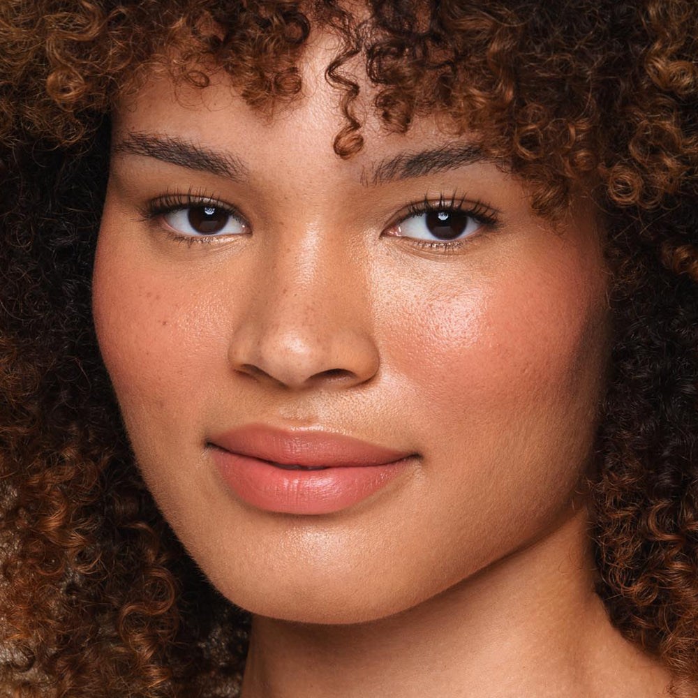 A close-up of a person with curly brown hair and glowing skin, smiling softly. The individual has full lips and well-defined eyebrows, wearing natural makeup that accentuates their features—achieved with the Essential Liquid Concealer from Jouer Cosmetics for a flawless, paraben-free finish.