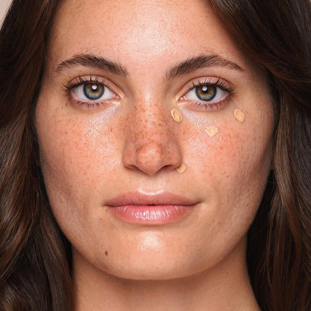 A close-up of a young woman with fair skin and freckles applying the *Essential Liquid Concealer* from Jouer Cosmetics under her eyes. She has green eyes, long brown hair, and slightly parted lips. The creamy, paraben-free concealer is dotted on her cheeks just below her eyes as she uses a concealer brush. The background is light and blurred.