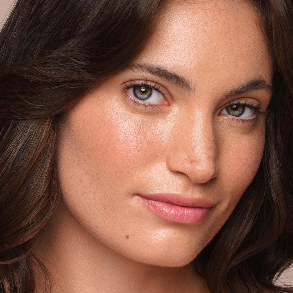 Close-up of a person's face with a serene expression. They have wavy, dark brown hair, light skin with a few freckles, hazel eyes, and are wearing a light pink lipstick. The lighting highlights their natural skin texture and facial features, subtly enhanced with the *Essential Liquid Concealer from Jouer Cosmetics for an effortless glow.
