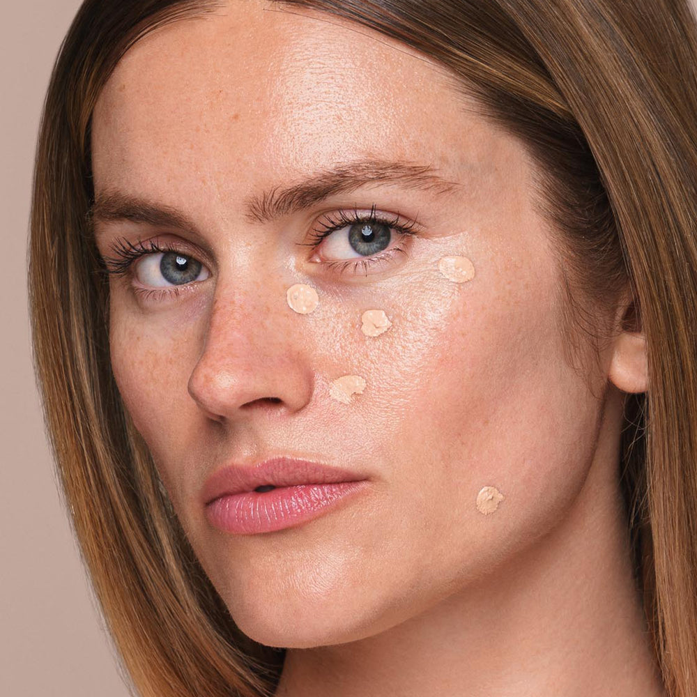 A woman with light skin and straight, dark blonde hair uses an *Essential Liquid Concealer* brush from Jouer Cosmetics to apply small dots of their paraben-free concealer to her face, focusing on her cheeks and under-eye area. She looks directly at the camera with a neutral expression.