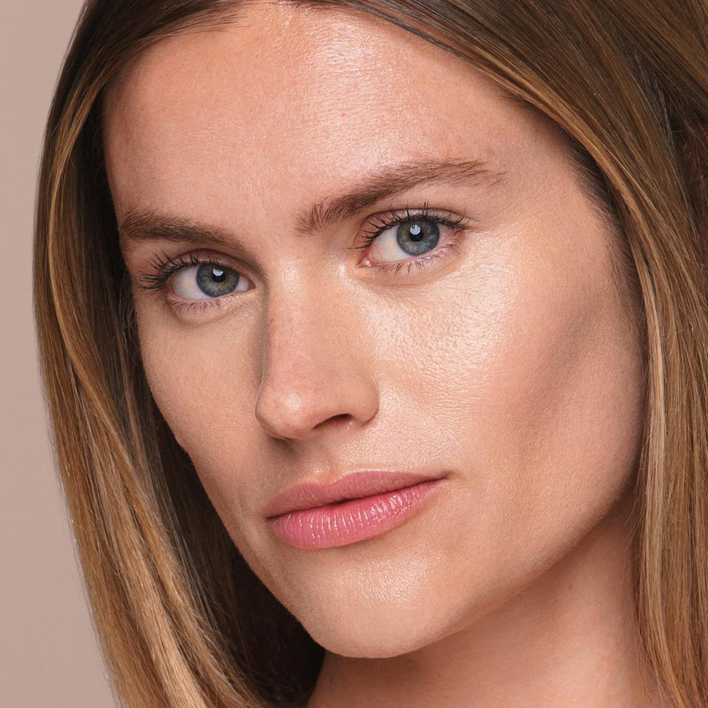 A close-up image features a person with straight, light brown hair. Their blue eyes and neutral expression stand out against a solid, neutral background. The lighting accentuates the person's clear skin and natural makeup, expertly applied using Jouer Cosmetics' *Essential Liquid Concealer for a flawless finish.