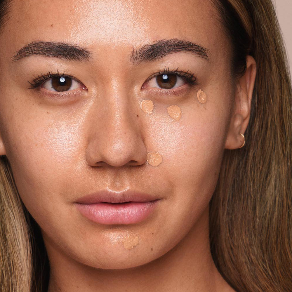 Close-up of a person with medium skin tone applying foundation dots to their face using an *Essential Liquid Concealer* brush by Jouer Cosmetics. The person has long hair, natural eyebrows, and minimal makeup, with visible foundation dots on their cheeks and chin. The background is neutral.