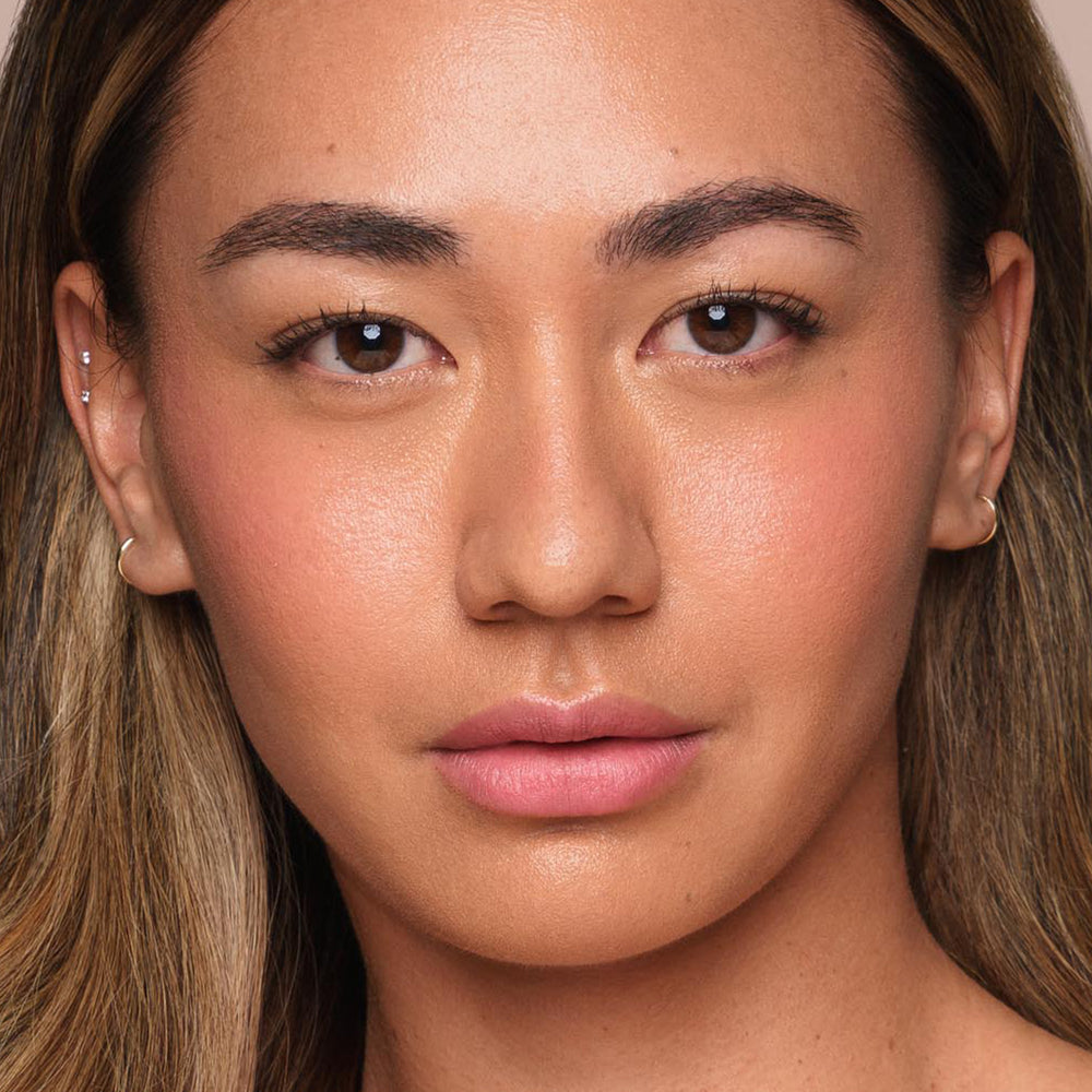 The image depicts a close-up of a person with long, straight, light brown hair and a clear complexion. They have brown eyes, neatly groomed eyebrows, and a neutral facial expression. Wearing subtle, vegan makeup from Jouer Cosmetics' *Essential Liquid Concealer*, they appear effortlessly flawless thanks to the natural look applied using the concealer brush.