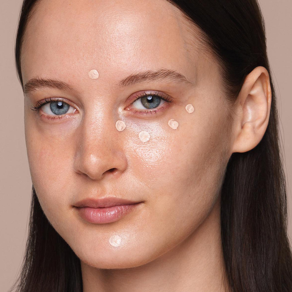 A person with light skin, blue eyes, and brown hair is looking directly at the camera. They have several small dots of Jouer Cosmetics' Essential Liquid Concealer, which is paraben-free, applied with a concealer brush to their face, including under their eyes, on their cheeks, and forehead. The background is neutral.
