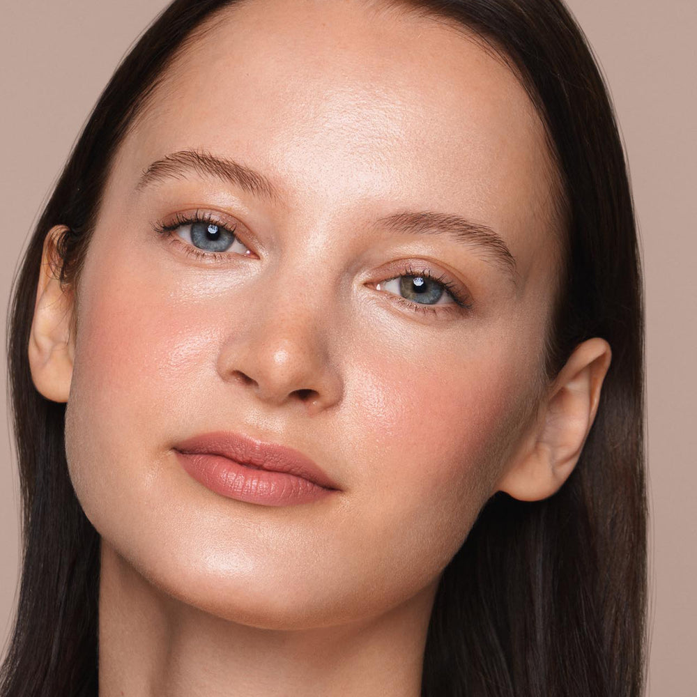 Close-up of a person with fair skin, light blue eyes, and dark brown hair. They have a neutral expression and are looking into the camera. The person is wearing natural-looking, vegan makeup including light blush, subtle eye makeup applied with Jouer Cosmetics' *Essential Liquid Concealer*, and a nude lip color. The background is plain.