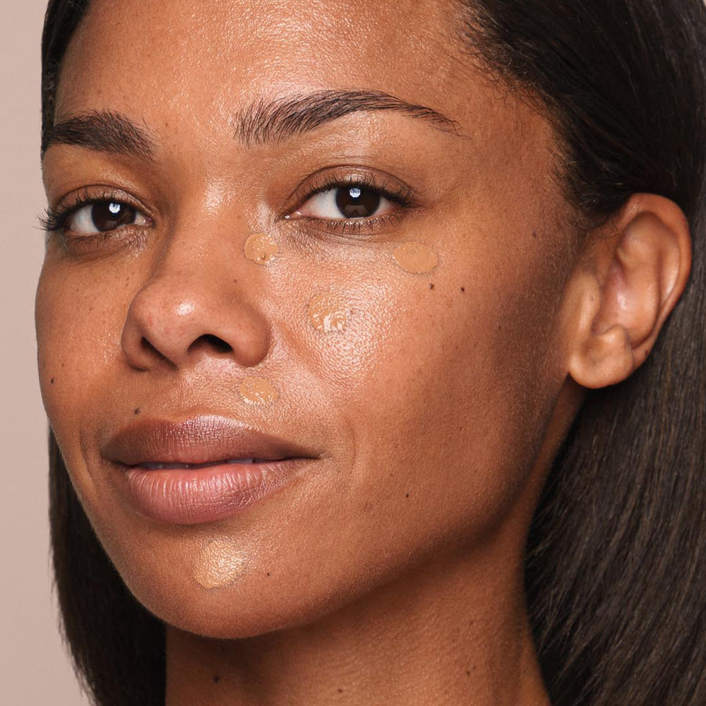 A woman with medium-dark skin and black hair looks directly at the camera. With a few drops of *Essential Liquid Concealer by Jouer Cosmetics applied to her cheeks and forehead, she is smiling slightly.