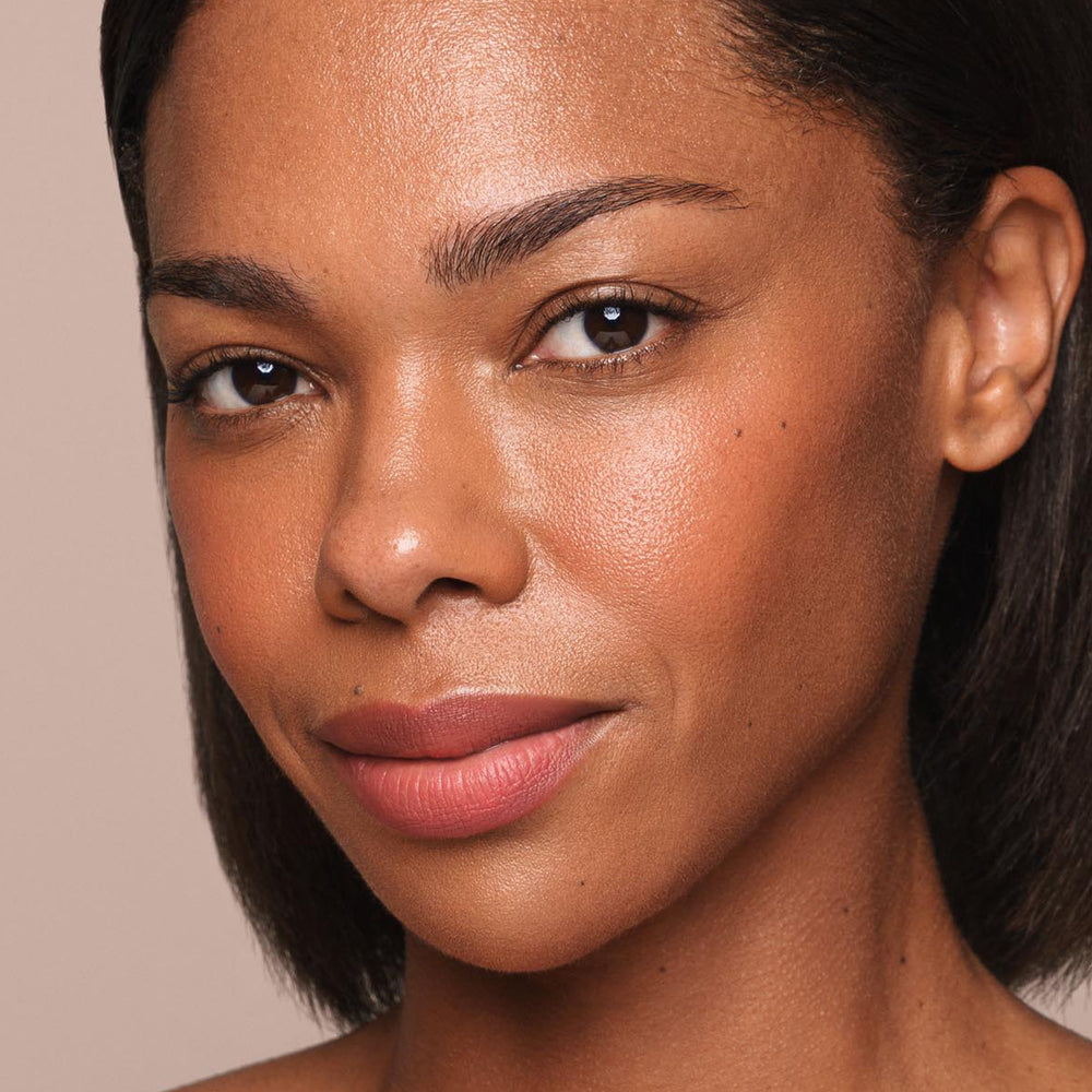 Close-up portrait of a woman with medium skin tone, short straight black hair, and well-defined eyebrows. She is looking directly at the camera with a slight smile, showcasing a natural makeup look featuring subtle blush and nude lipstick applied using Jouer Cosmetics' *Essential Liquid Concealer*. The background is plain beige.
