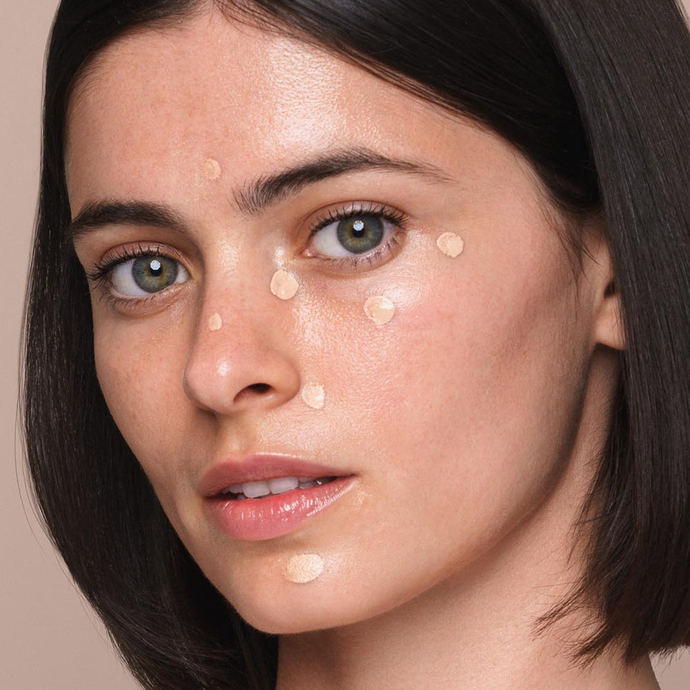 A close-up of a woman with medium-length dark hair, light skin, and green eyes. She has small dots of the *Essential Liquid Concealer* by Jouer Cosmetics applied on various points of her face, including her forehead, cheeks, nose, and chin. A neutral background highlights the focus on her radiant complexion.