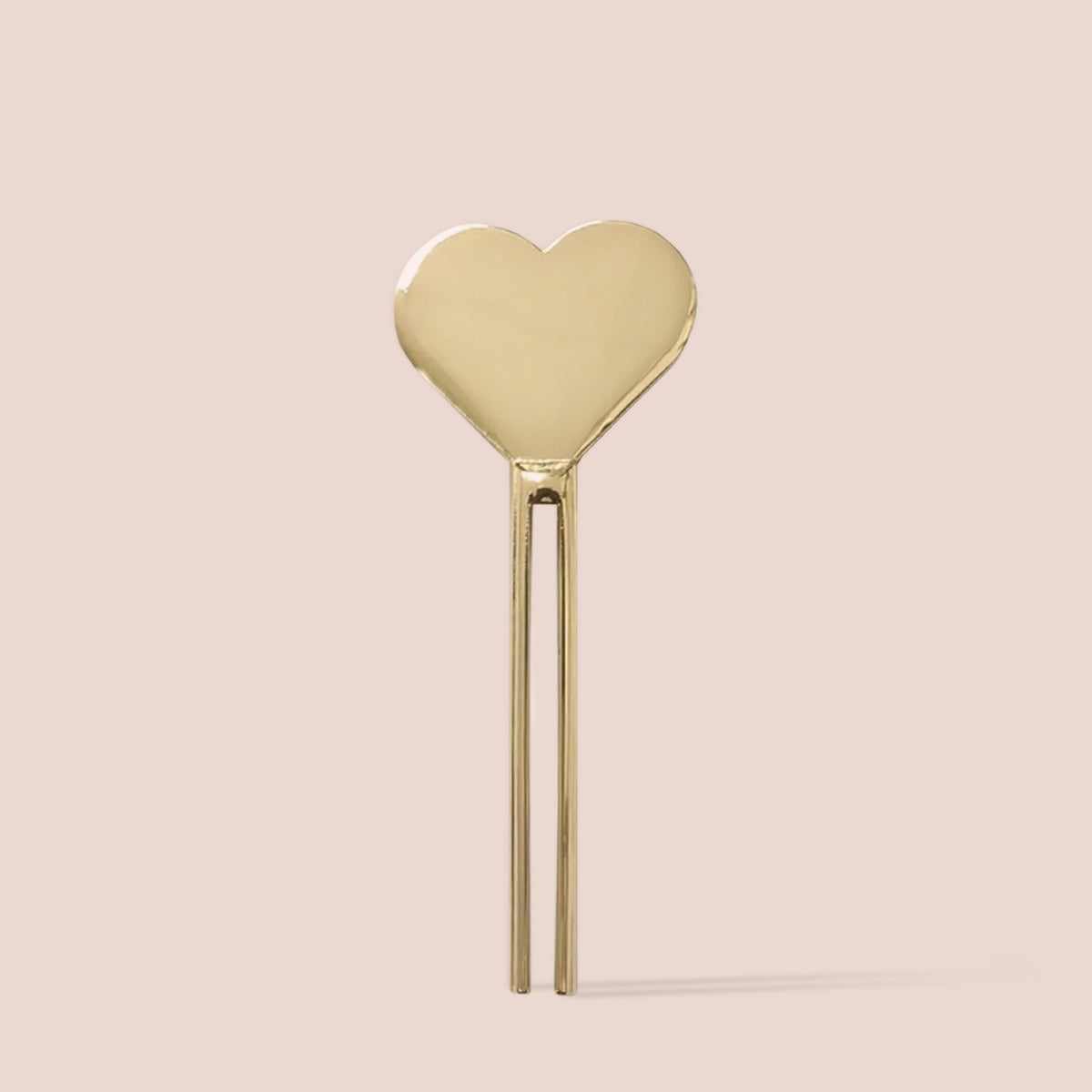 Gold heart-shaped metal tube key on a soft blush background, designed for squeezing cosmetic tubes.
