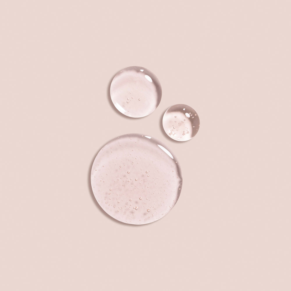 Three transparent droplets of Jouer Cosmetics' Essential Lip Oil are scattered on a smooth beige surface, arranged with one large droplet at the bottom and two smaller droplets positioned above it. This minimalist and aesthetically pleasing composition embodies a gentle, paraben-free essence suitable for any vegan lifestyle.