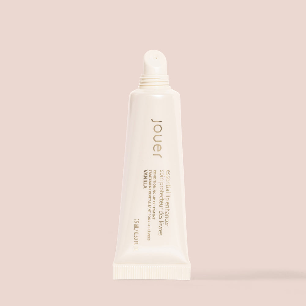 A tube of Jouer Cosmetics Essential Lip Enhancer, which is both cruelty-free and paraben-free, stands upright on a beige background. The off-white tube with a narrow nozzle displays text that identifies it as a conditioning lip treatment in vanilla flavor, containing 0.5 fl oz or 15 mL.
